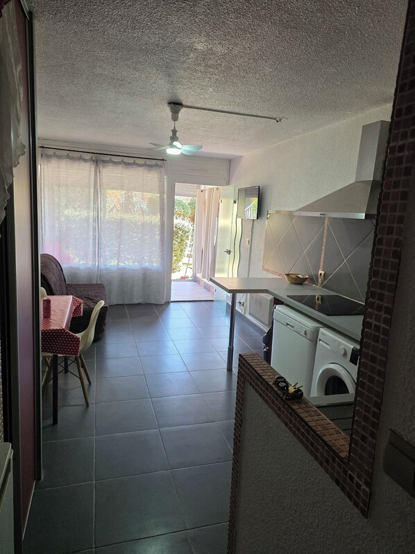 A compact kitchen with a washing machine, a dining area with a table and chairs, and a view of the outdoors through a door.