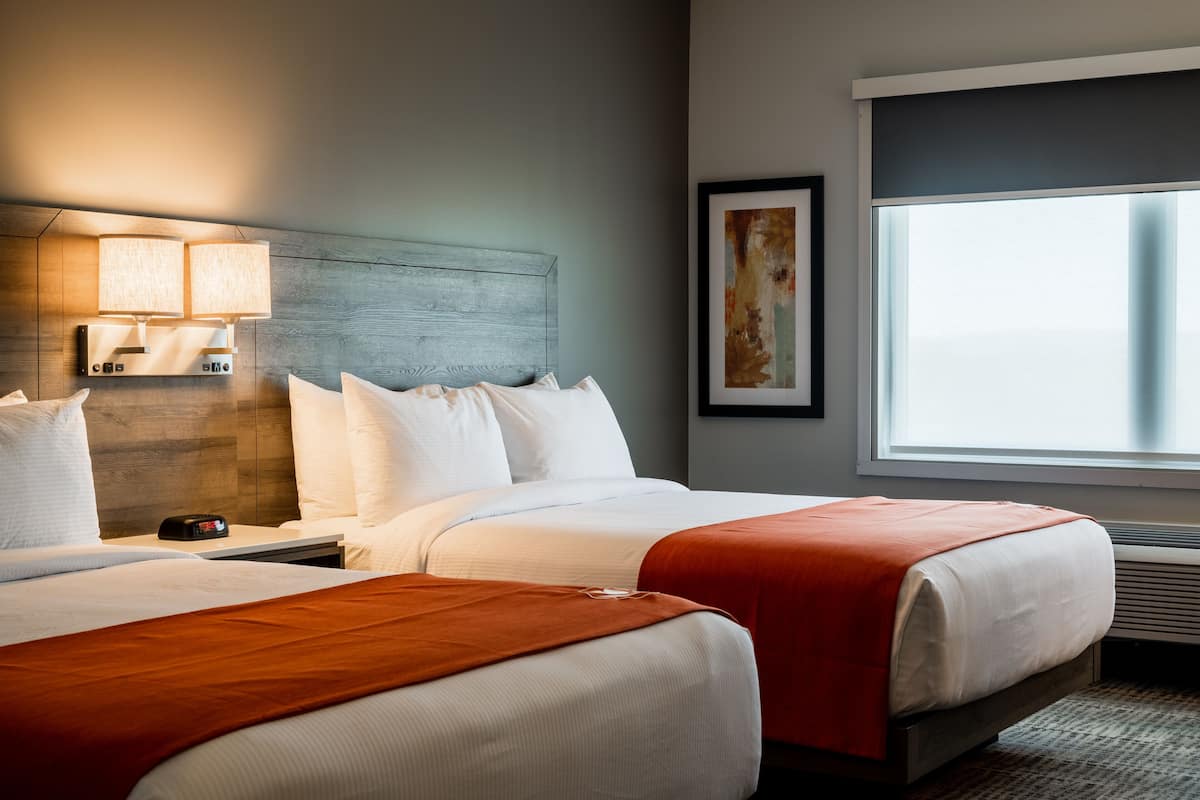 Een hotelkamer met twee bedden, een nachtkastje met een lamp, een kunstwerk in een lijst aan de muur en een groot raam met uitzicht.