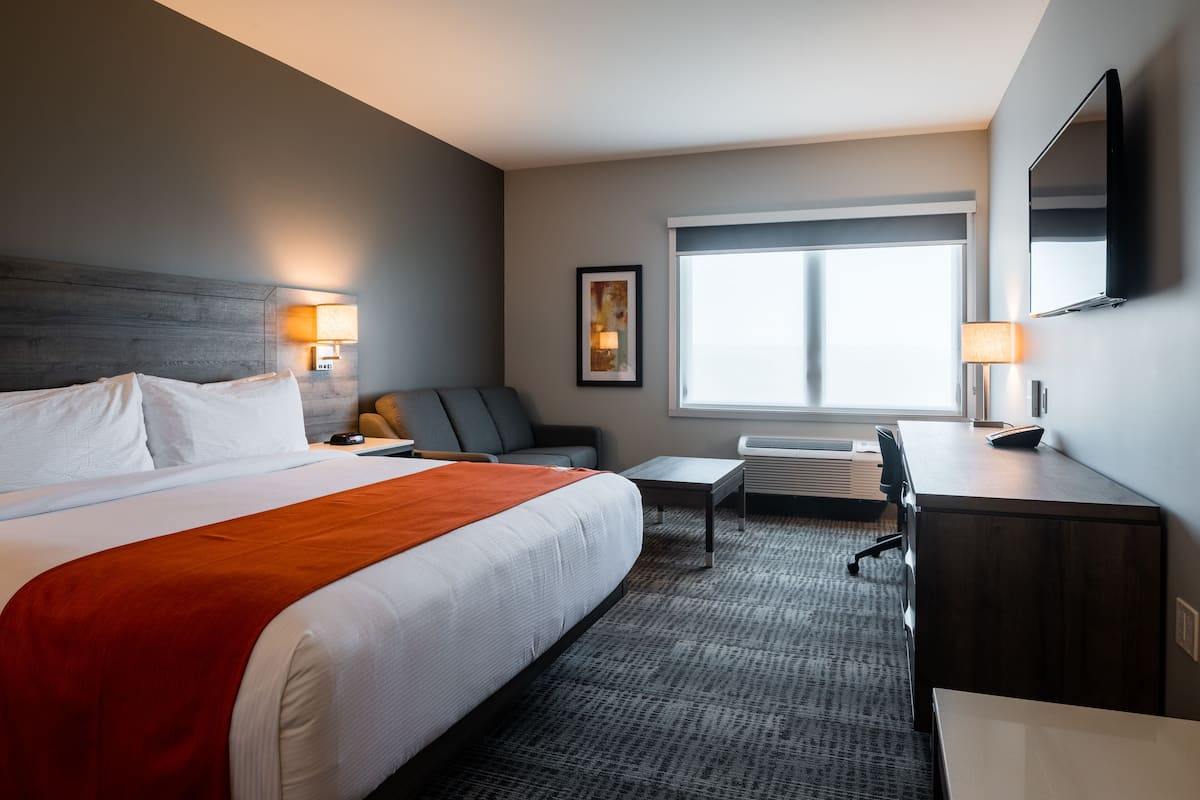 Een hotelkamer met een groot bed, een bureau, een stoel, een kleine tafel, een lamp en een raam met zonwering.