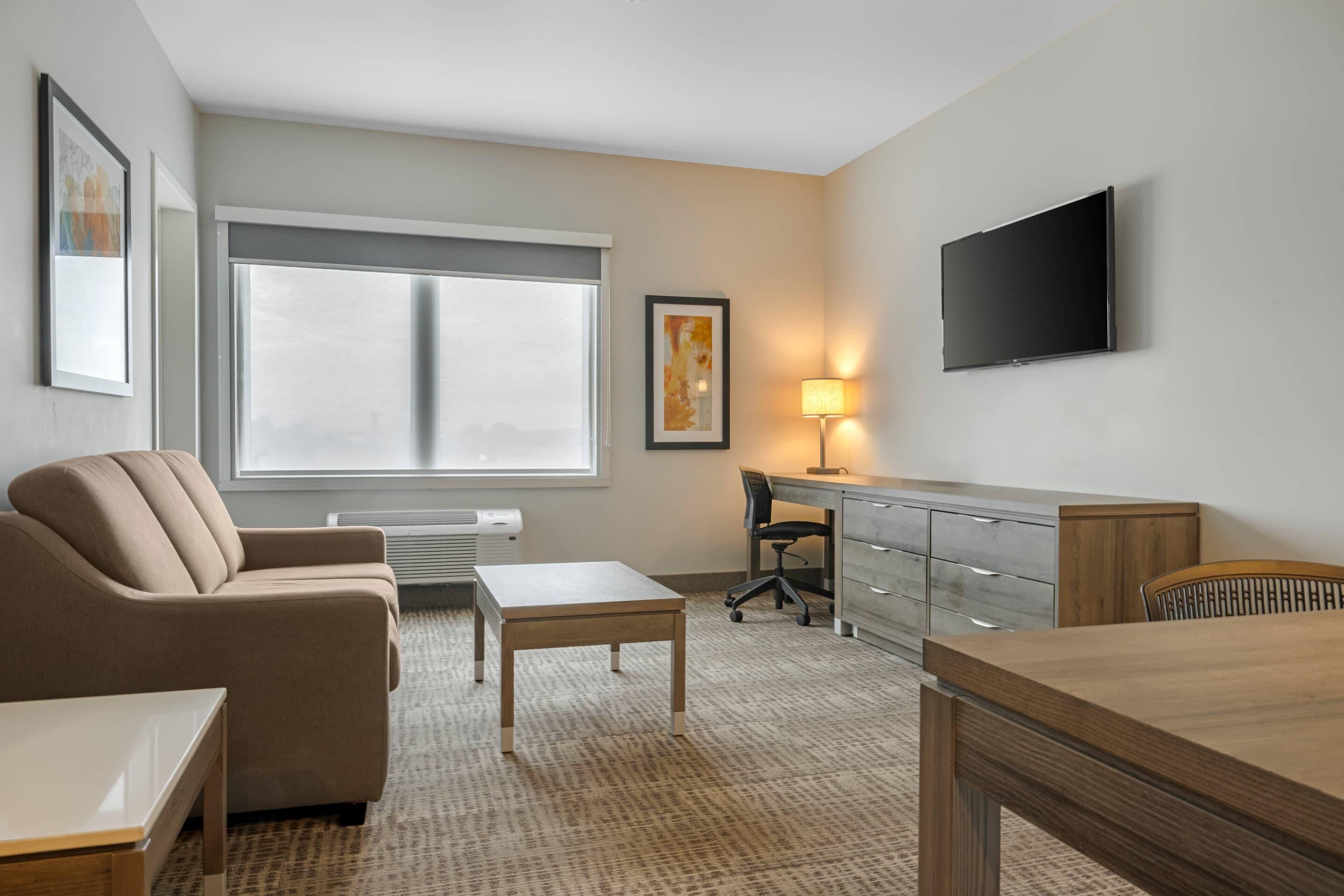 A hotel room with a sofa, a desk, a TV, a lamp, and a large window.