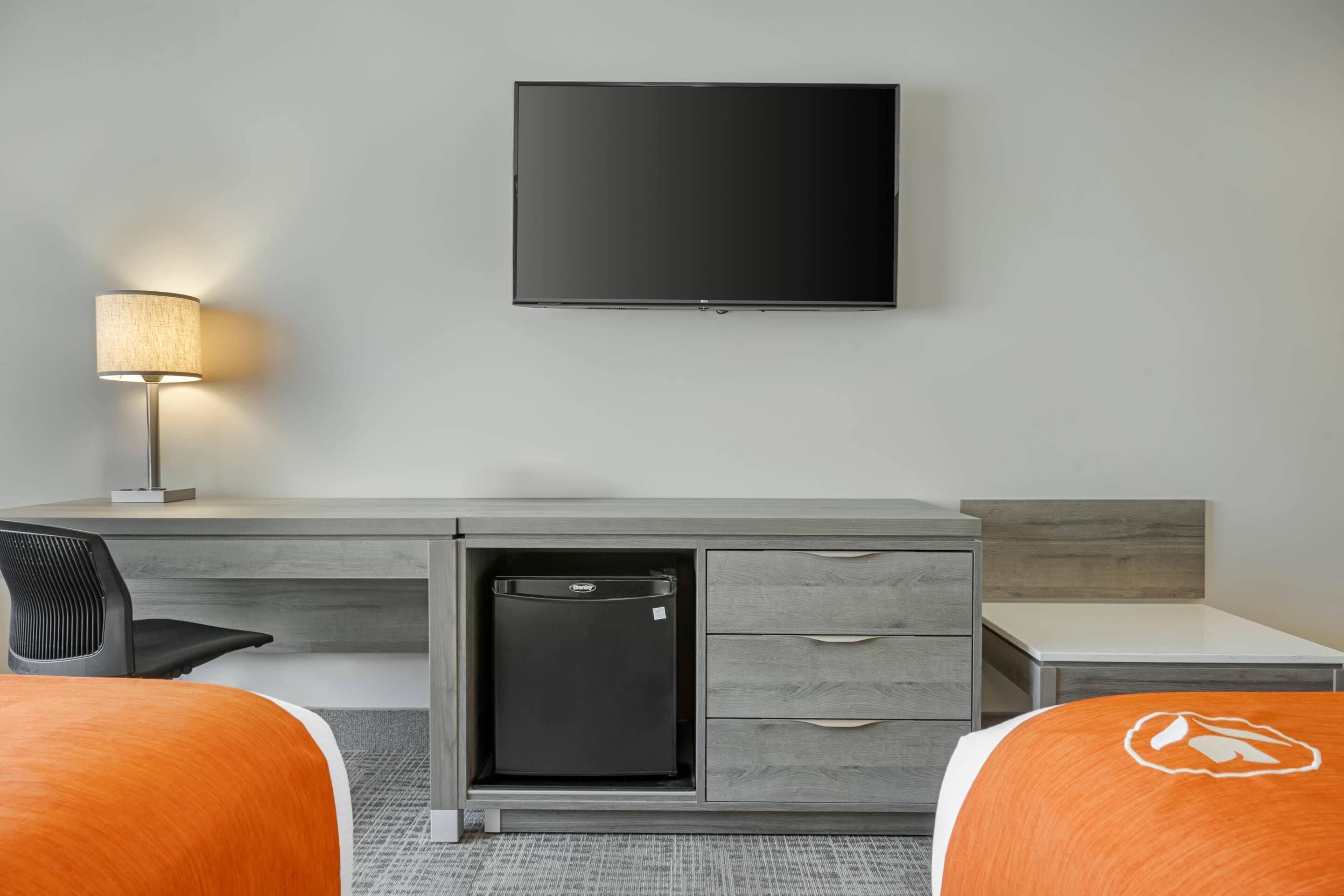 A hotel room with a flat-screen TV mounted on the wall, a desk with a lamp, a mini fridge, and two beds with orange bedding.