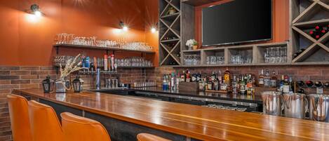 A bar area with a long counter, orange bar stools, shelves with glasses and bottles, and a large TV screen.