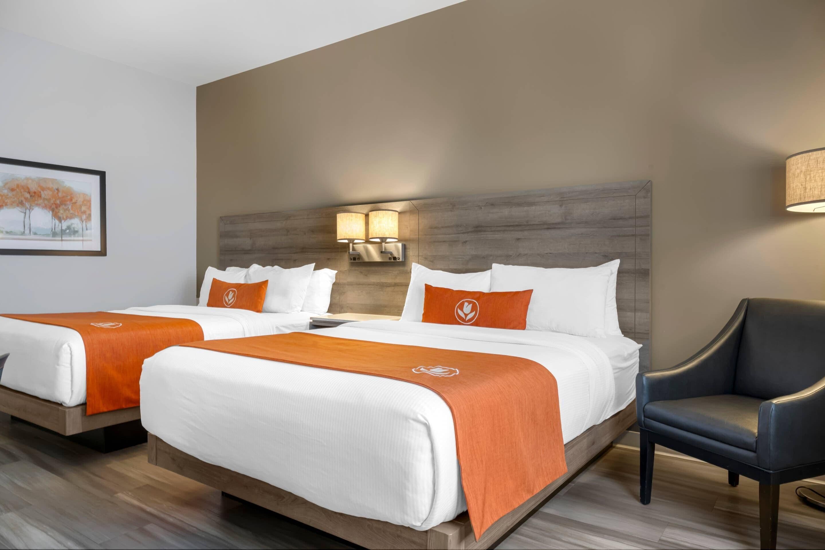 Habitación de hotel con dos camas, cada una con ropa blanca y detalles naranja, cabeceros de madera y una lámpara fijada a la pared.