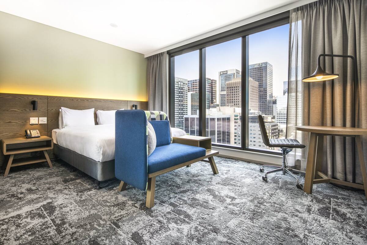 Una habitación de hotel con un ventanal grande, una cama, una silla azul, un escritorio y una lámpara.