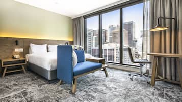 Quarto de hotel com cama, poltrona azul, escrivaninha, cadeira, luminária e uma grande janela com vista para a cidade.
