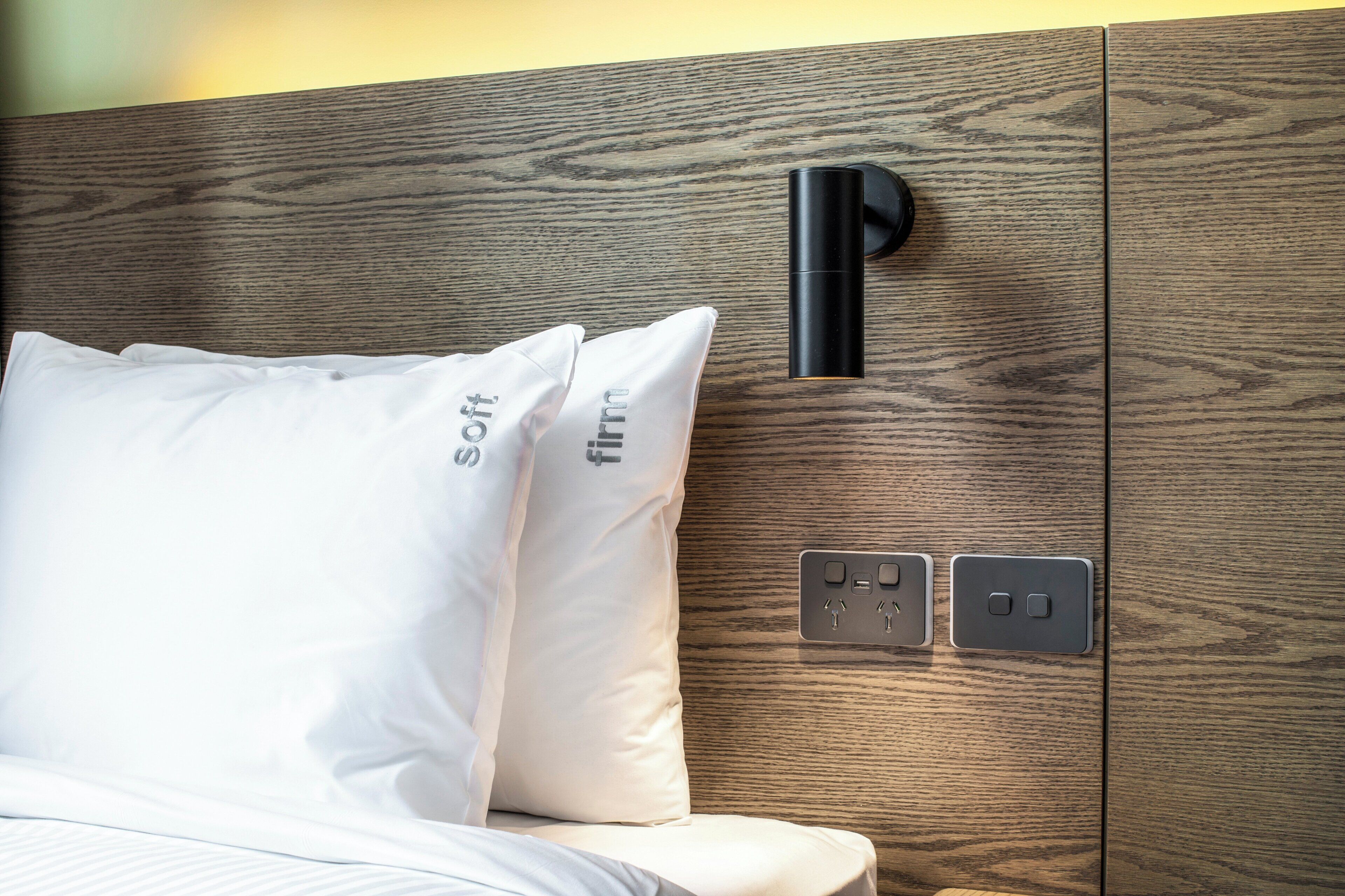 Un lit avec une literie et des oreillers blancs, une tête de lit en bois, une lampe noire fixée au mur et une prise de courant dotée de ports USB.