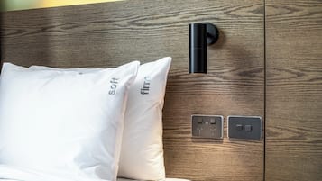 Un lit avec une literie et des oreillers blancs, une tête de lit en bois, une lampe noire fixée au mur et une prise de courant dotée de ports USB.
