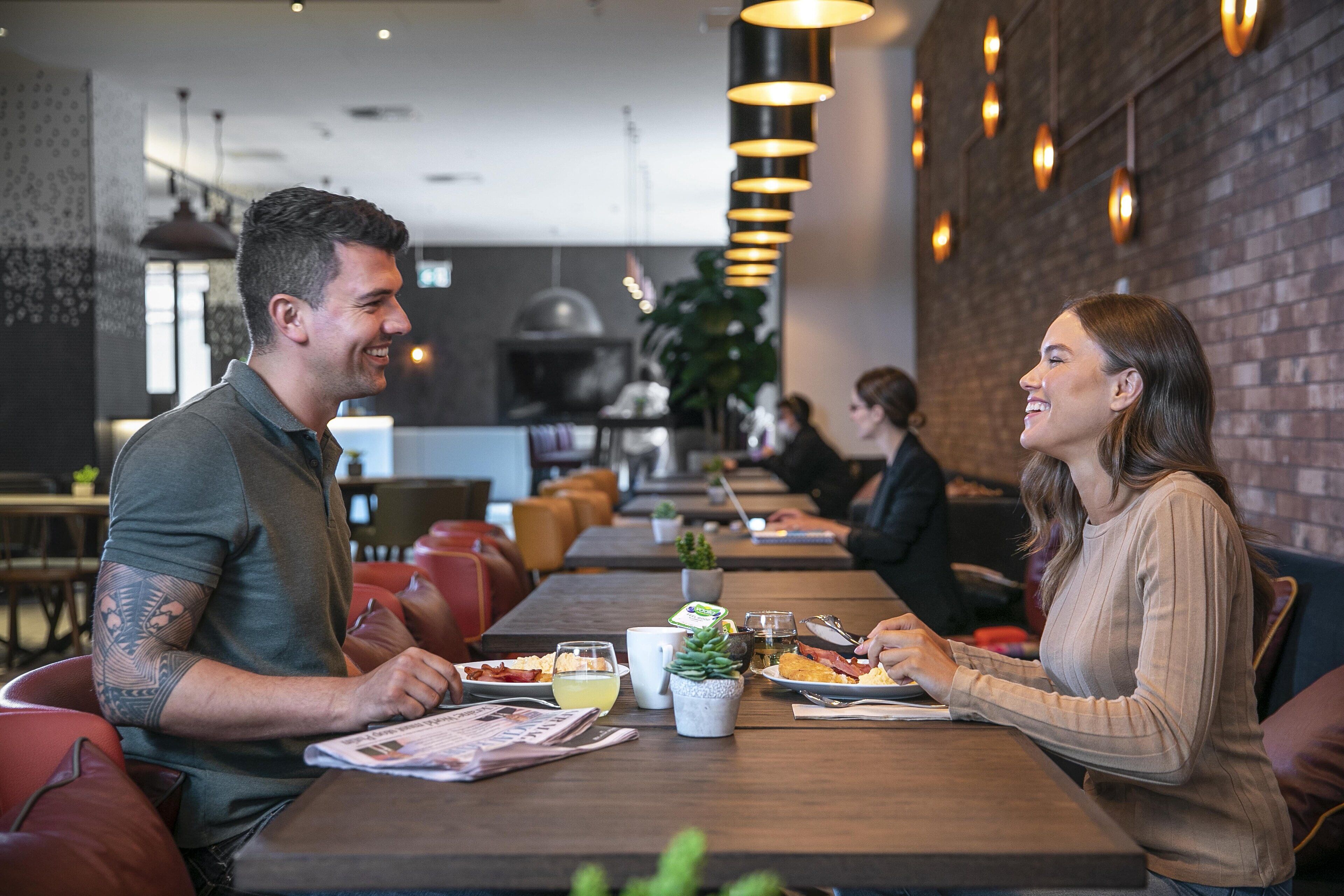 Restaurante com mesas e cadeiras de madeira, parede de tijolos e luminárias pendentes.