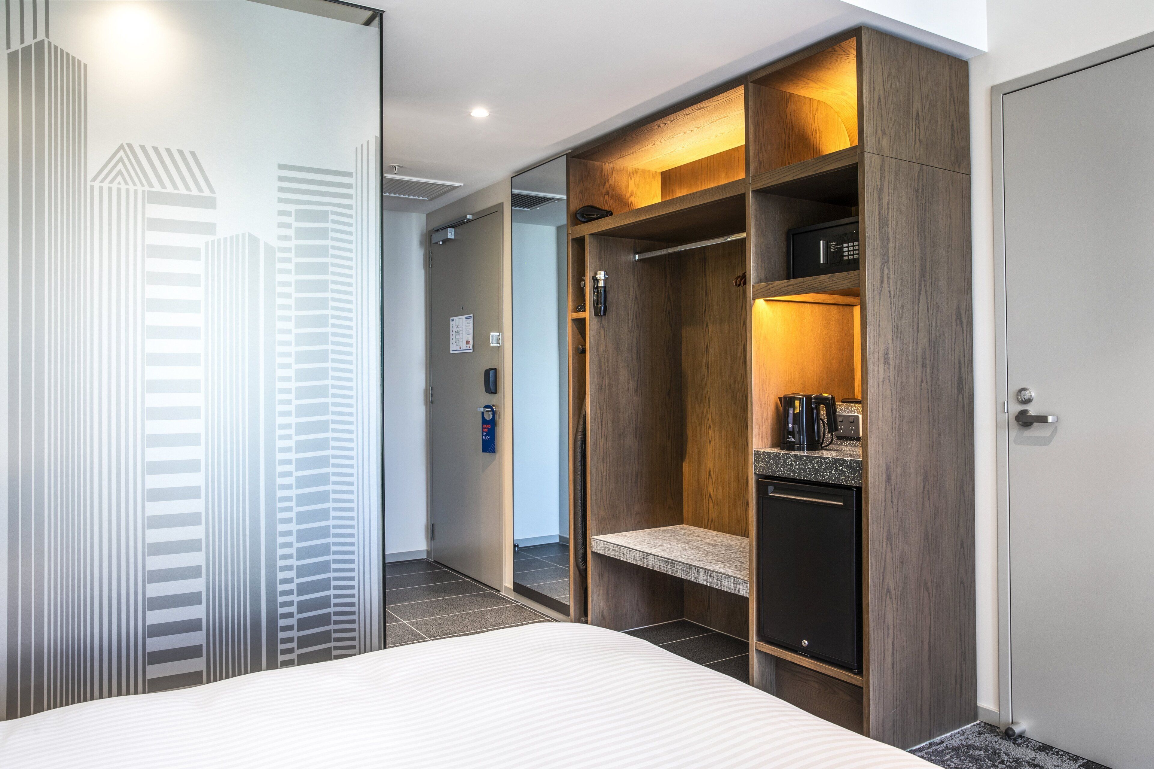 Une chambre d’hôtel avec un lit, une armoire en bois, une petite kitchenette et une vue sur de hauts immeubles.