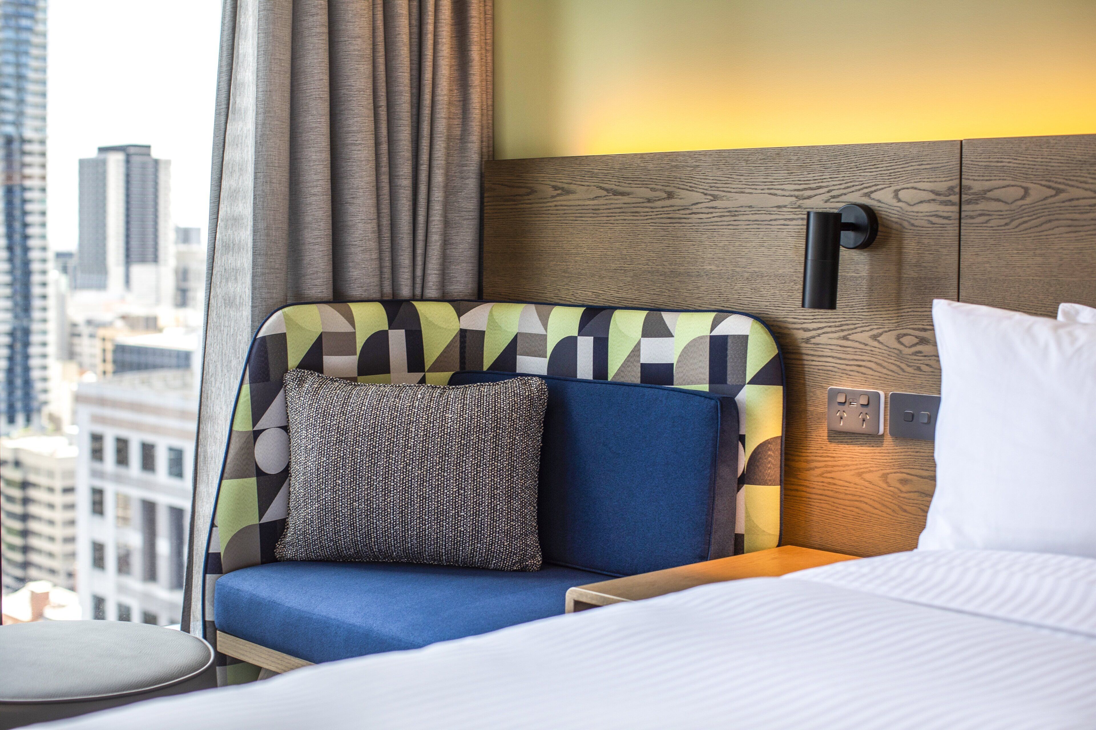 A hotel room with a bed, a seating area with a cushion, a small table, and a view of the cityscape through large windows.
