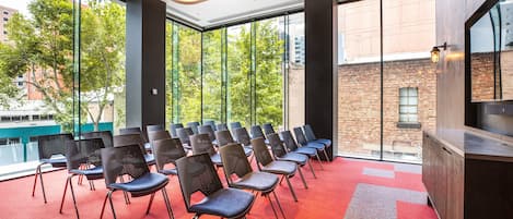 Uma sala de conferências com fileiras de cadeiras, janelas amplas e uma parede de tijolos visível através do vidro.