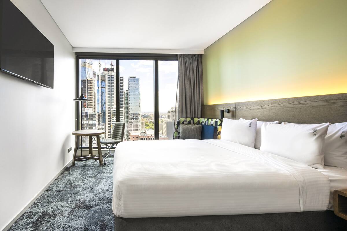 Habitación de hotel con una cama grande, un escritorio, una silla y vistas a la ciudad a través de la ventana.