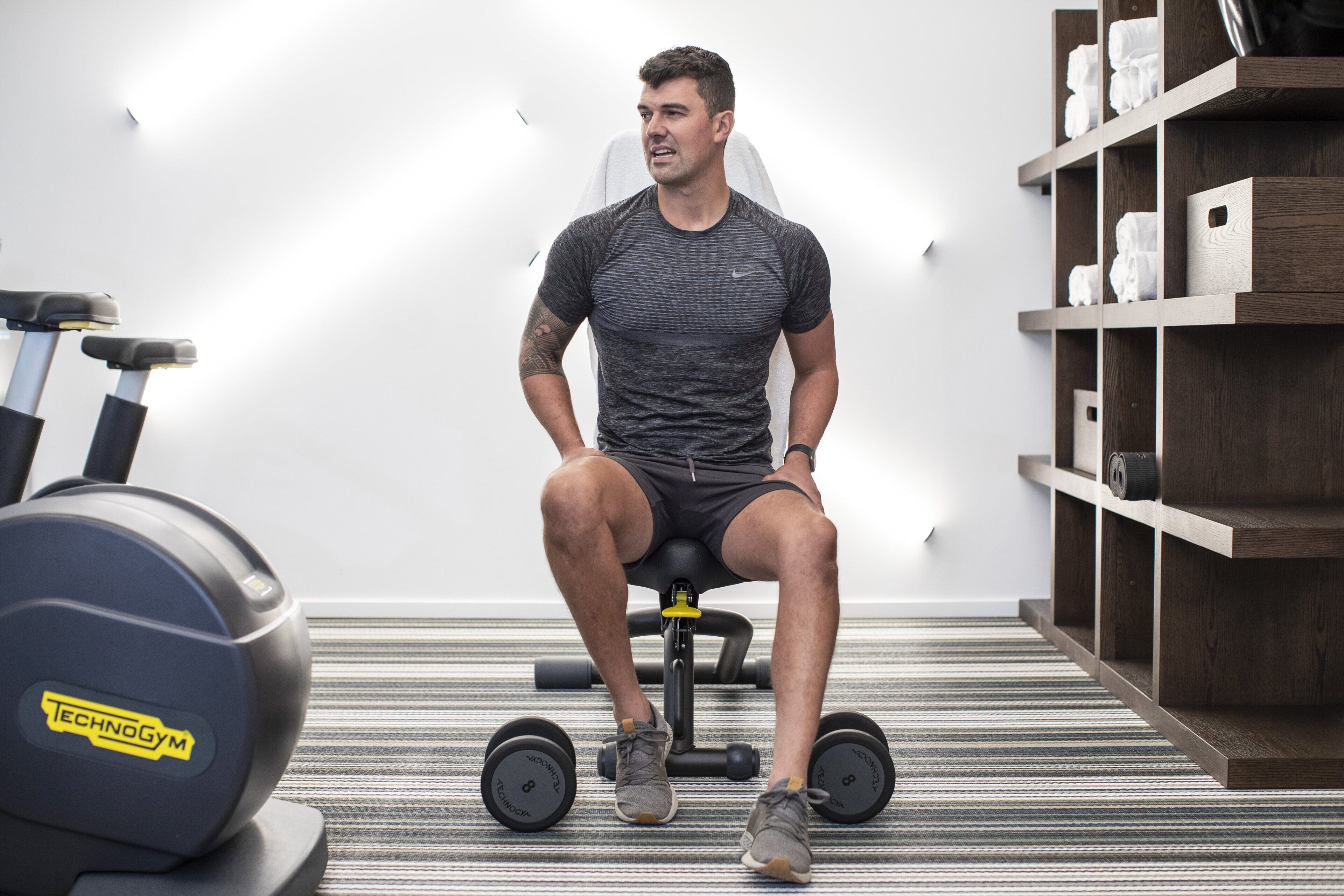 A person seated on a stationary bike in a room with a striped carpet, a shelf with towels, and a wall-mounted light.