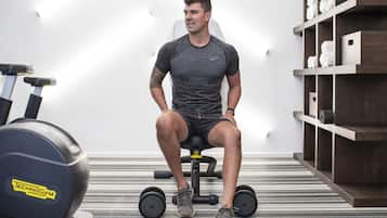 A person seated on a stationary bike in a room with a striped carpet, a shelf with towels, and a wall-mounted light.