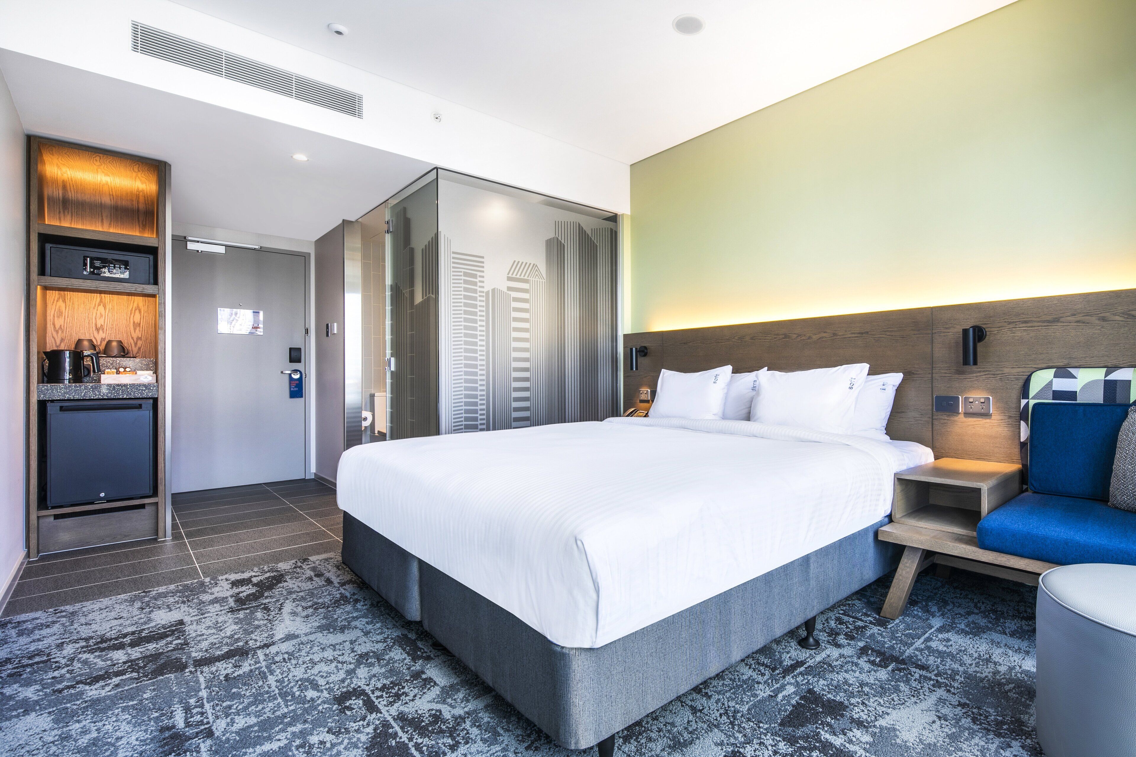 A modern hotel room with a large bed, a seating area with a blue sofa, a small table, and a built-in kitchen area with a coffee machine.