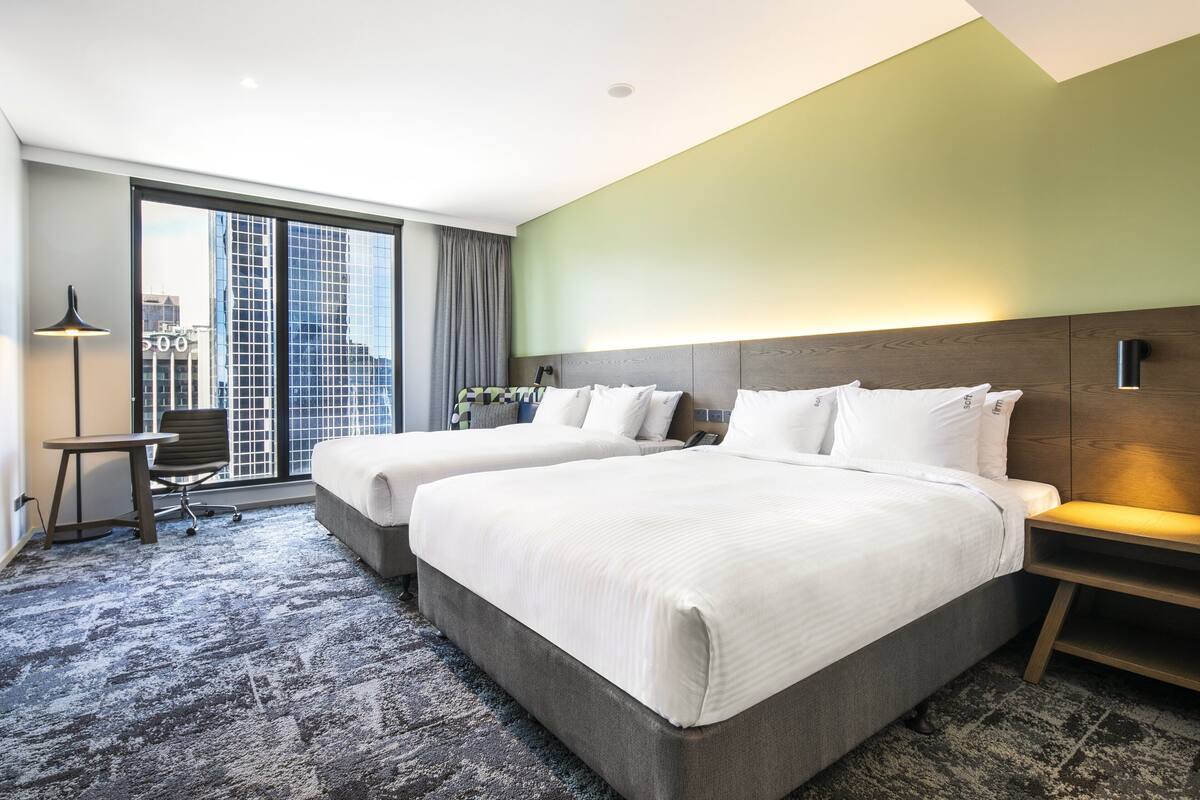 Habitación de hotel con dos camas, un escritorio, una silla y un ventanal con vistas a la ciudad.