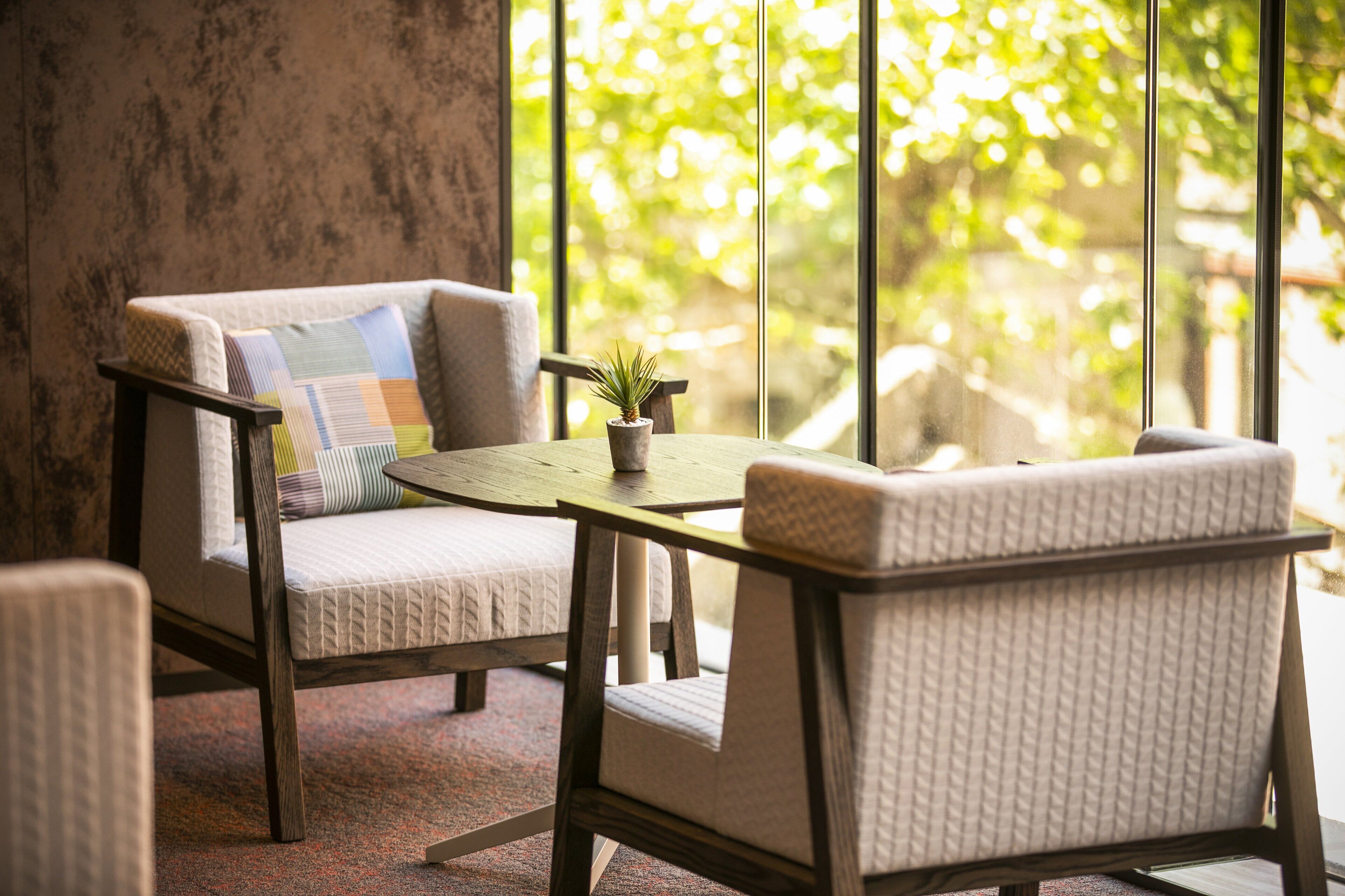 Un rincón acogedor con un sillón con cojines, una mesita pequeña, una planta en maceta y vistas a la vegetación a través de grandes ventanales.