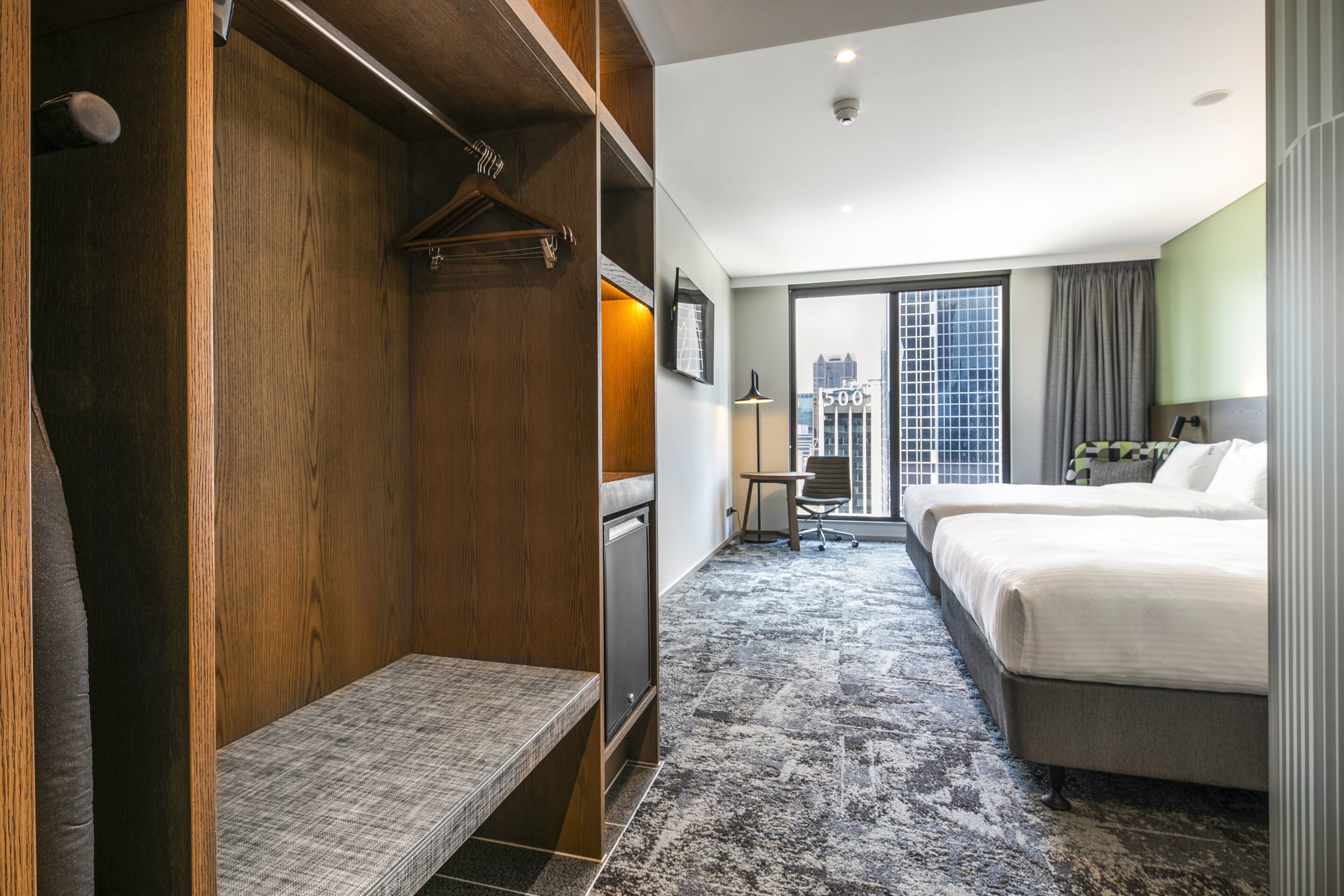 Een hotelkamer met een houten garderobe, een bankje, twee bedden, een bureau en een groot raam met uitzicht op de stad.