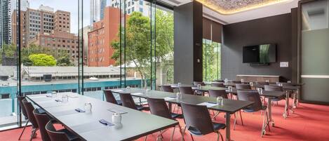 A conference room with tables and chairs, large windows with a city view, a flat-screen TV on the wall, and a ceiling with a unique pattern.