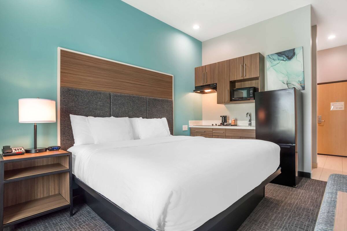 A hotel room with a large bed, a nightstand with a lamp, a microwave, and a refrigerator.