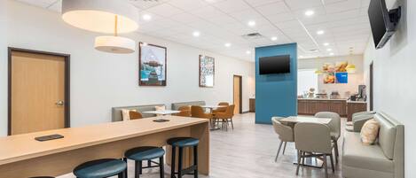 A modern hotel lobby with a long reception desk, multiple seating areas, and a kitchen in the background.