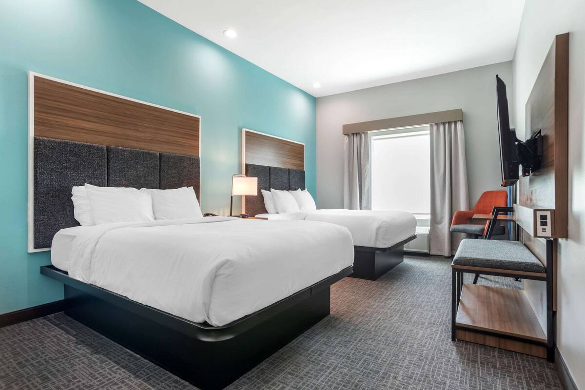 A hotel room with two beds, a TV mounted on the wall, a small table, and a window with curtains.