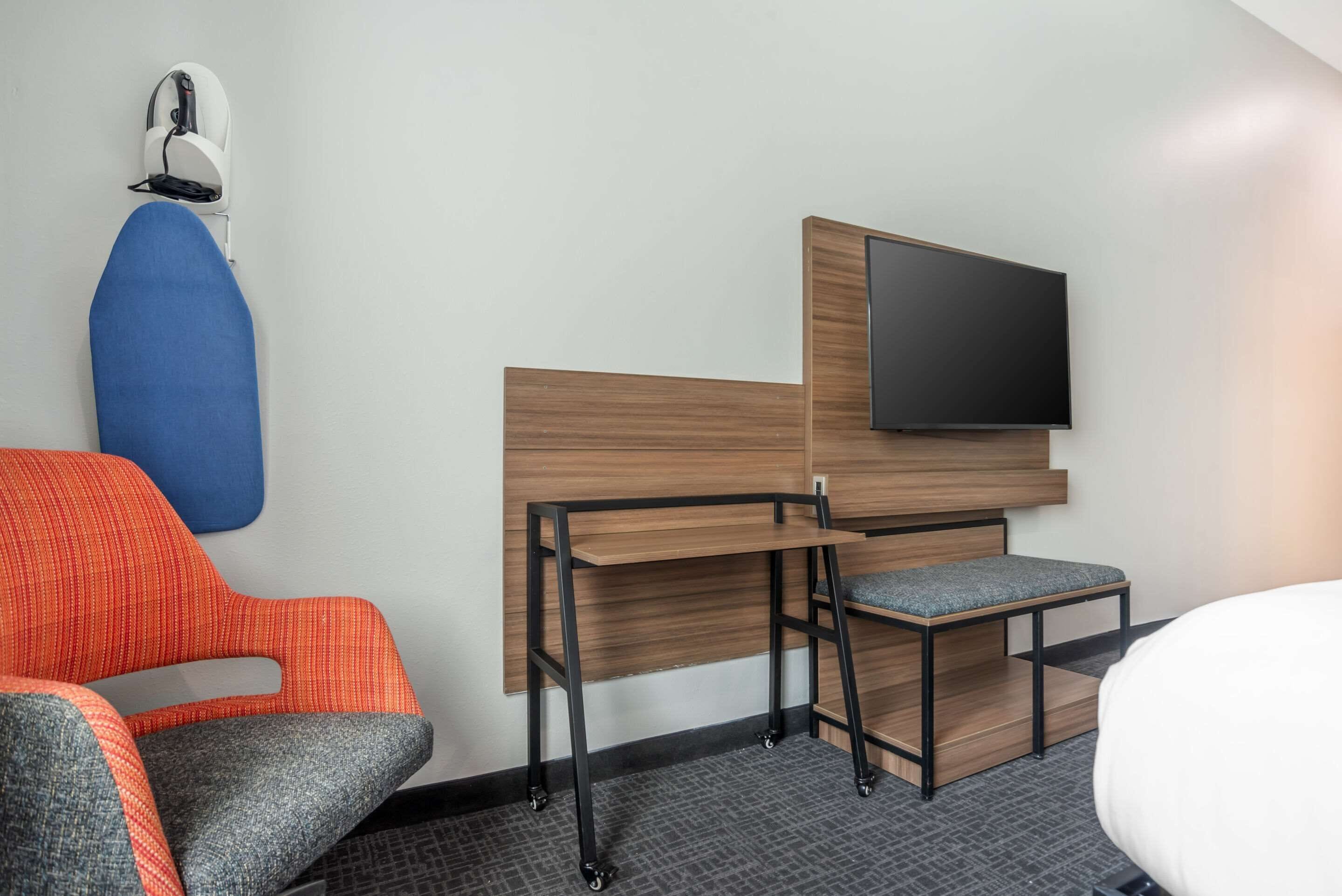 A hotel room with a bed, a desk with a chair, a flat-screen TV mounted on a wooden panel, and a blue and orange chair.