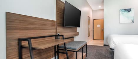 Habitación de hotel con cabecera de madera, televisor de pantalla plana montado encima, un pequeño banco con cojín, cama con ropa blanca y suelo alfombrado.