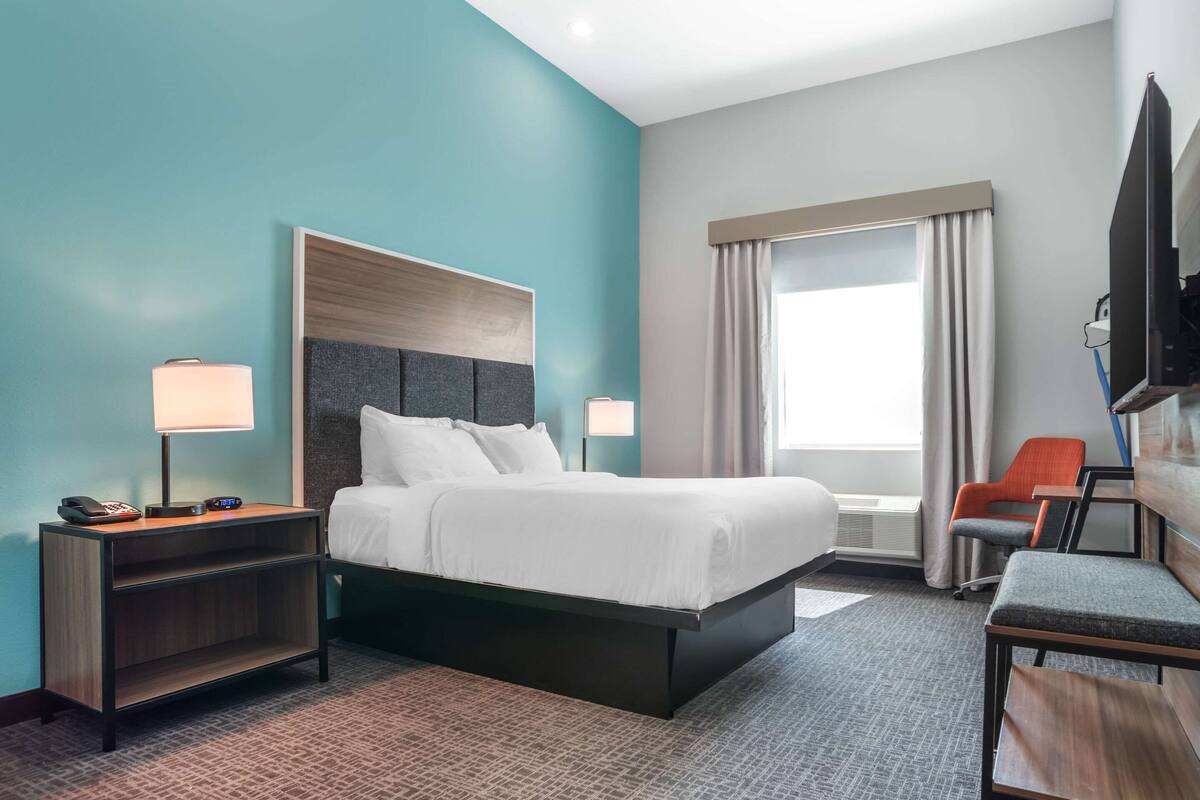 A hotel room with a bed, bedside tables, a desk, a chair, a TV, and a window with curtains.