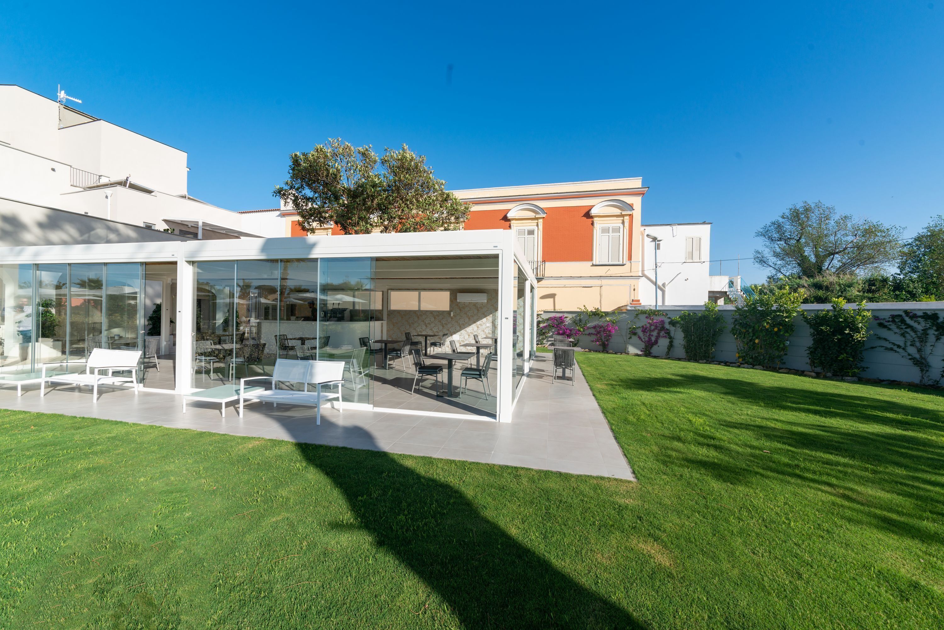 Un patio moderno al aire libre con paredes de vidrio, mobiliario blanco y un césped bien cuidado.