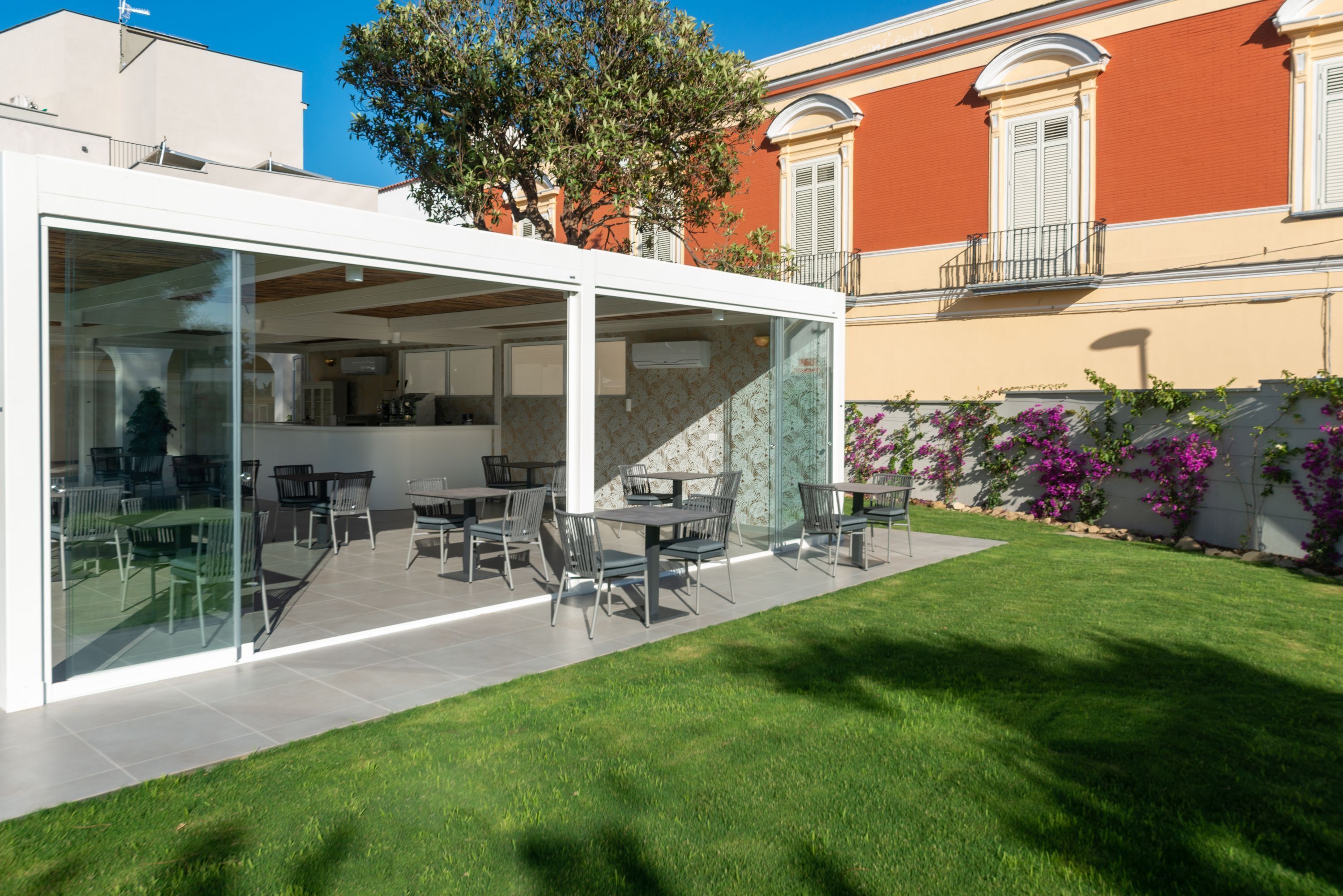 A modern outdoor dining area with glass walls, a neatly trimmed lawn, and a building with a balcony in the background.