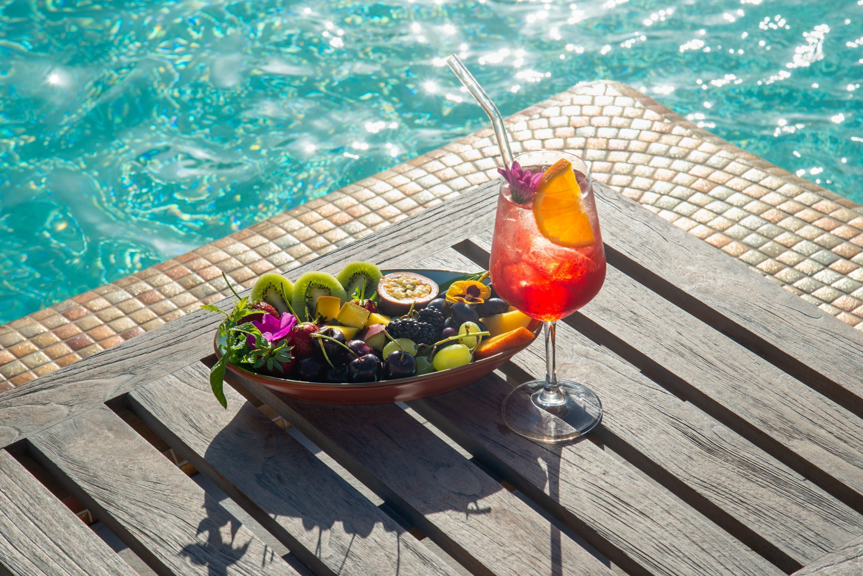 A wooden table with a fruit bowl and a cocktail glass beside a pool.