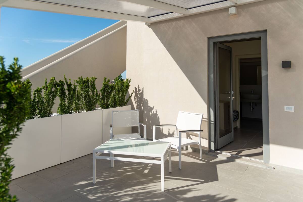 Un patio moderno al aire libre con una mesa de vidrio y sillas blancas, una pared blanca y una puerta que da a un espacio interior.