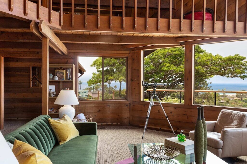 A wooden cabin interior with a green sofa, a tripod, and a view of the ocean.