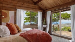 Une chambre avec un grand lit, des oreillers blancs et orange, une couverture rouge et une vue sur l’extérieur grâce à de grandes fenêtres.