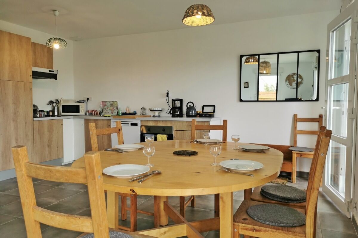 Un coin repas avec une table ronde en bois, dressée pour quatre personnes, entourée de chaises en bois dotées d’assises rembourrées. On aperçoit en arrière-plan une cuisine équipée d’appareils modernes et de meubles en bois.