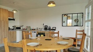 Un coin repas avec une table ronde en bois, dressée pour quatre personnes, entourée de chaises en bois dotées d’assises rembourrées. On aperçoit en arrière-plan une cuisine équipée d’appareils modernes et de meubles en bois.