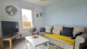Un salon avec un téléviseur, une table blanche, un canapé beige avec des coussins, une chaise en bois et une grande fenêtre.