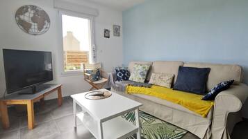 Un salon avec un téléviseur, une table blanche, un canapé beige avec des coussins, une chaise en bois et une grande fenêtre.