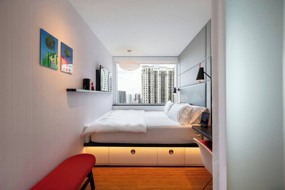 Un dormitorio moderno con cama, mesita de noche y ventana con vista a la ciudad.
