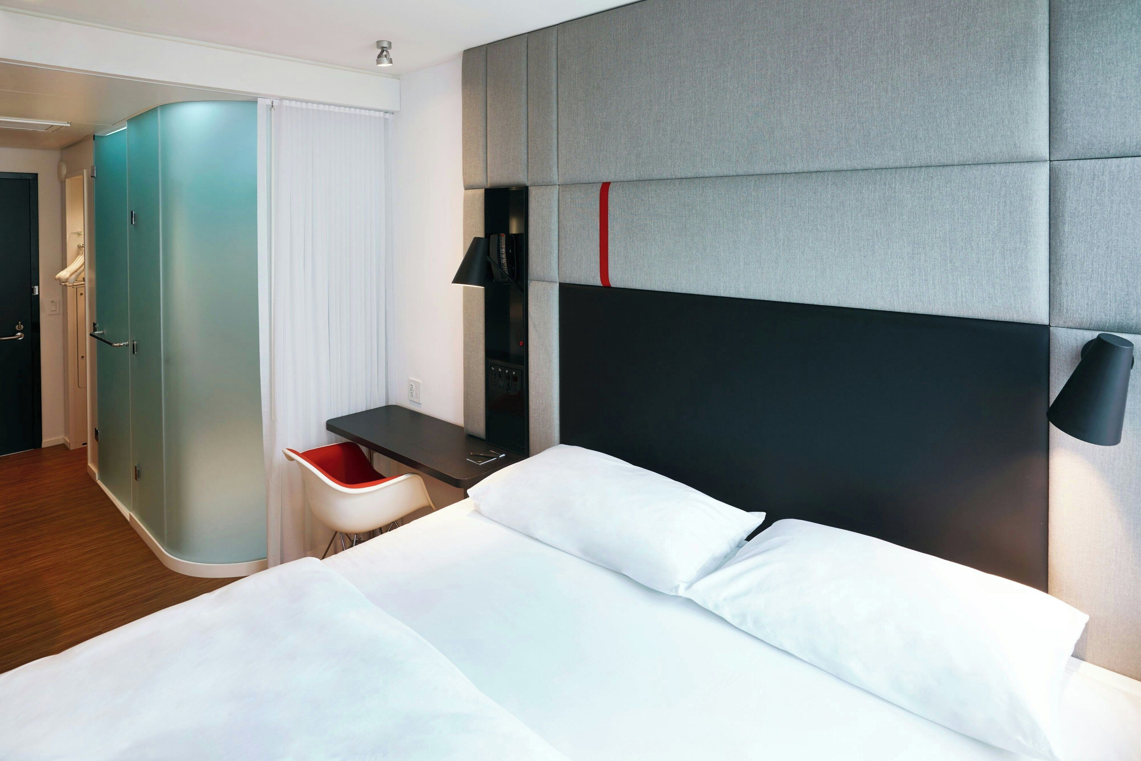 A hotel room with a bed, a desk, a chair, a wardrobe, and a wall-mounted lamp.