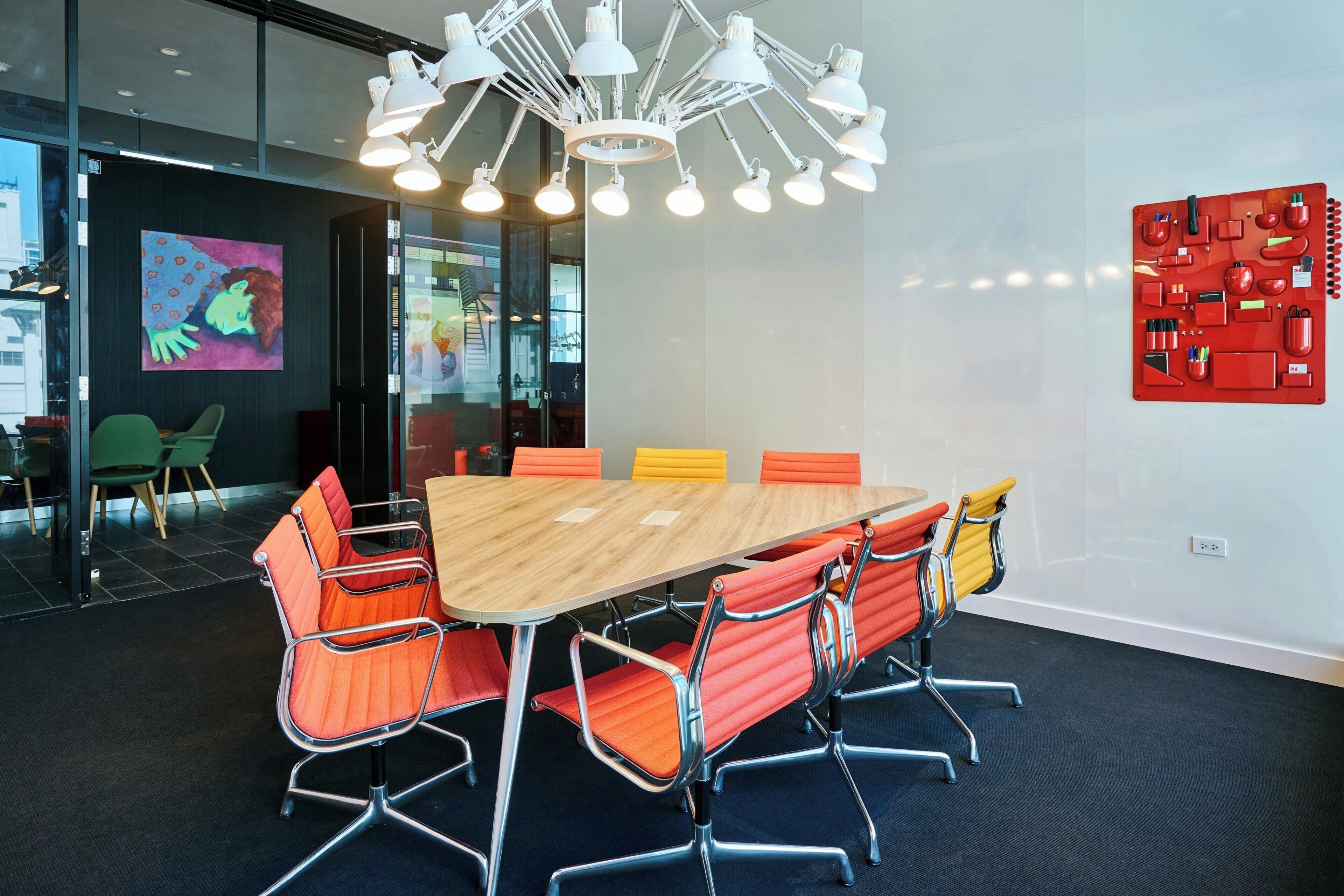 A conference room with a large table, orange chairs, a modern chandelier, and a colorful abstract painting on the wall.