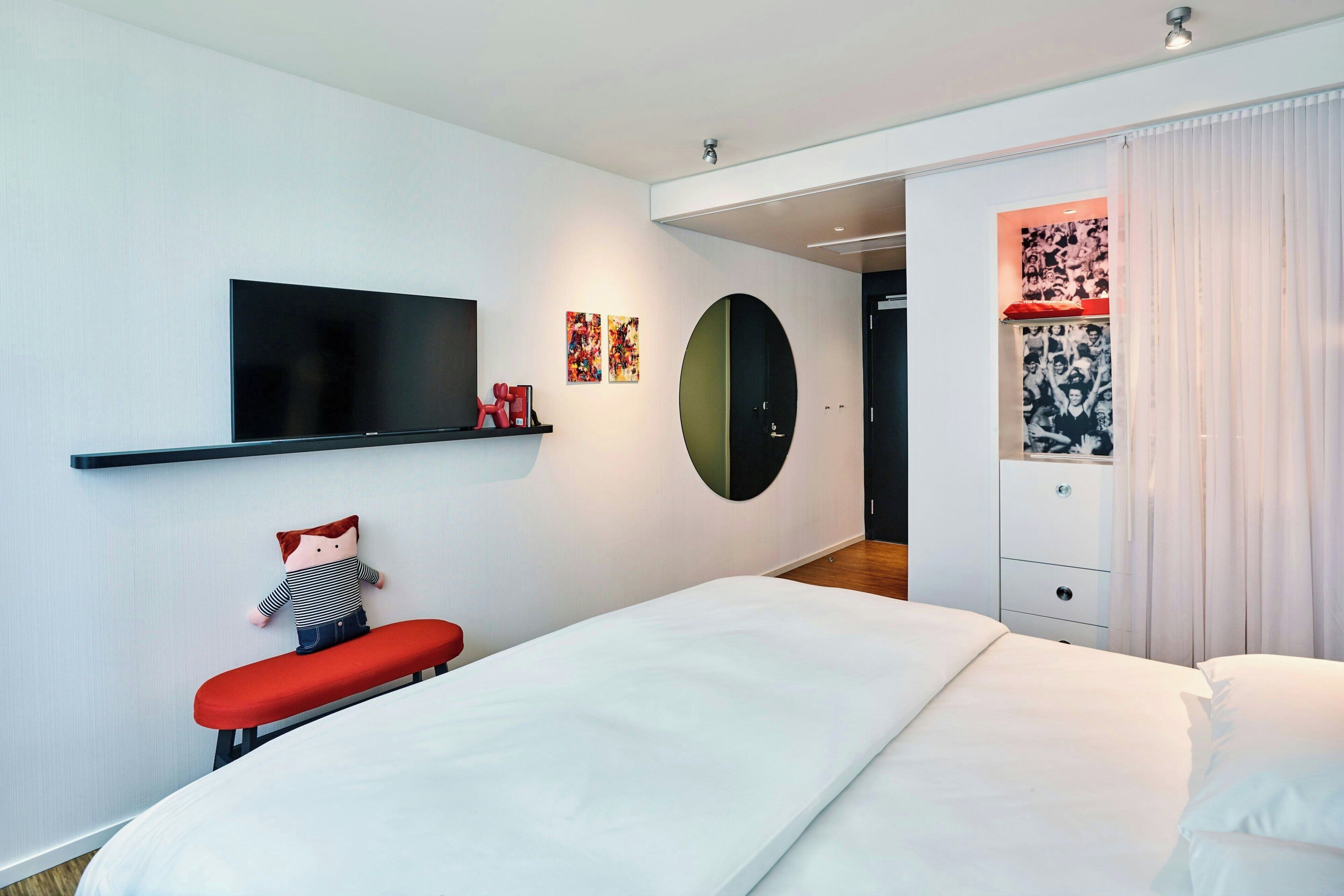 Une chambre d’hôtel moderne avec un grand lit, un téléviseur à écran plat, un banc rouge et une armoire avec un miroir.