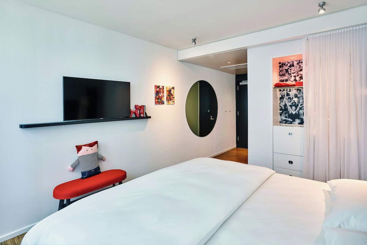 Ett modernt hotellrum med en stor säng, en platt-TV, en röd bänk och en garderob med en spegel.