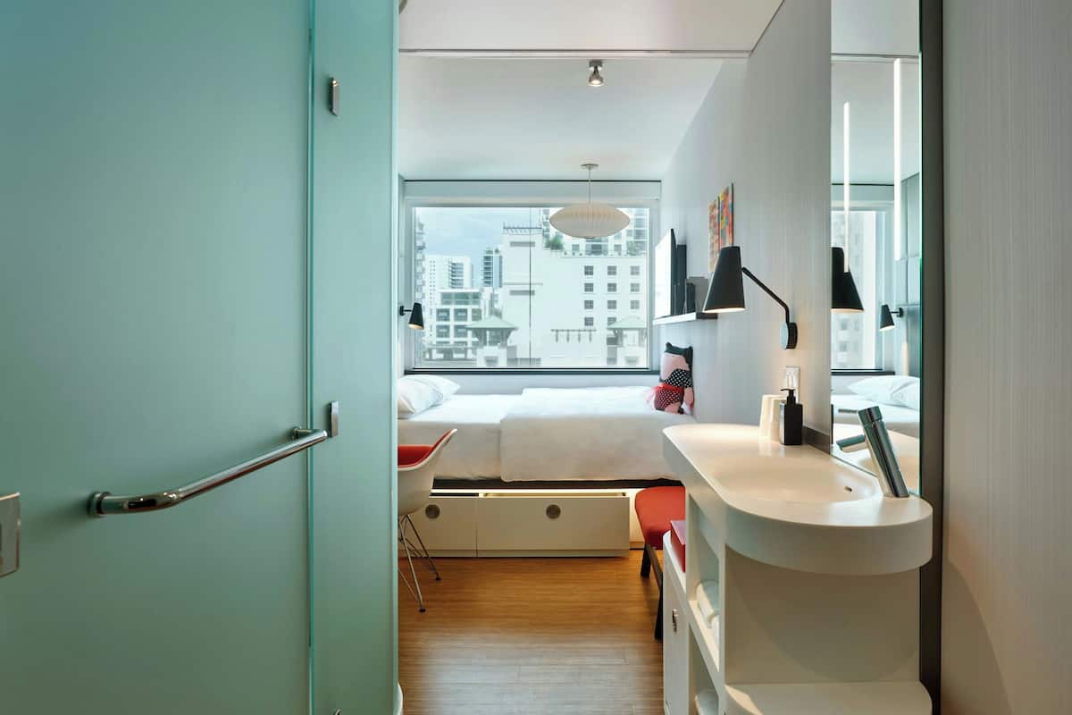 Un dormitorio moderno con un ventanal grande, una mesita de noche, una cama y un baño con lavamanos y espejo.