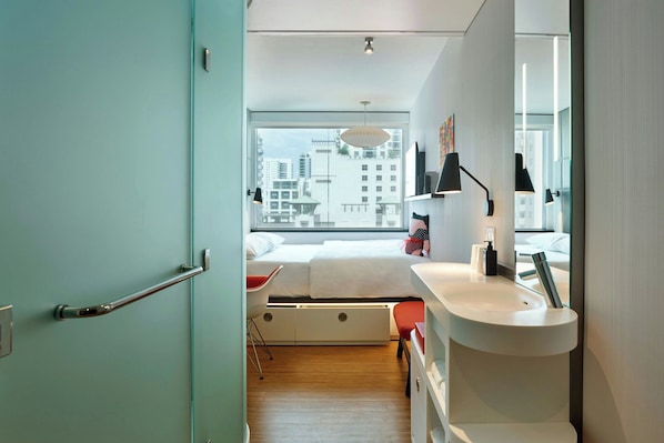 A modern bedroom with a large window, a bedside table, a bed, and a bathroom with a sink and mirror.