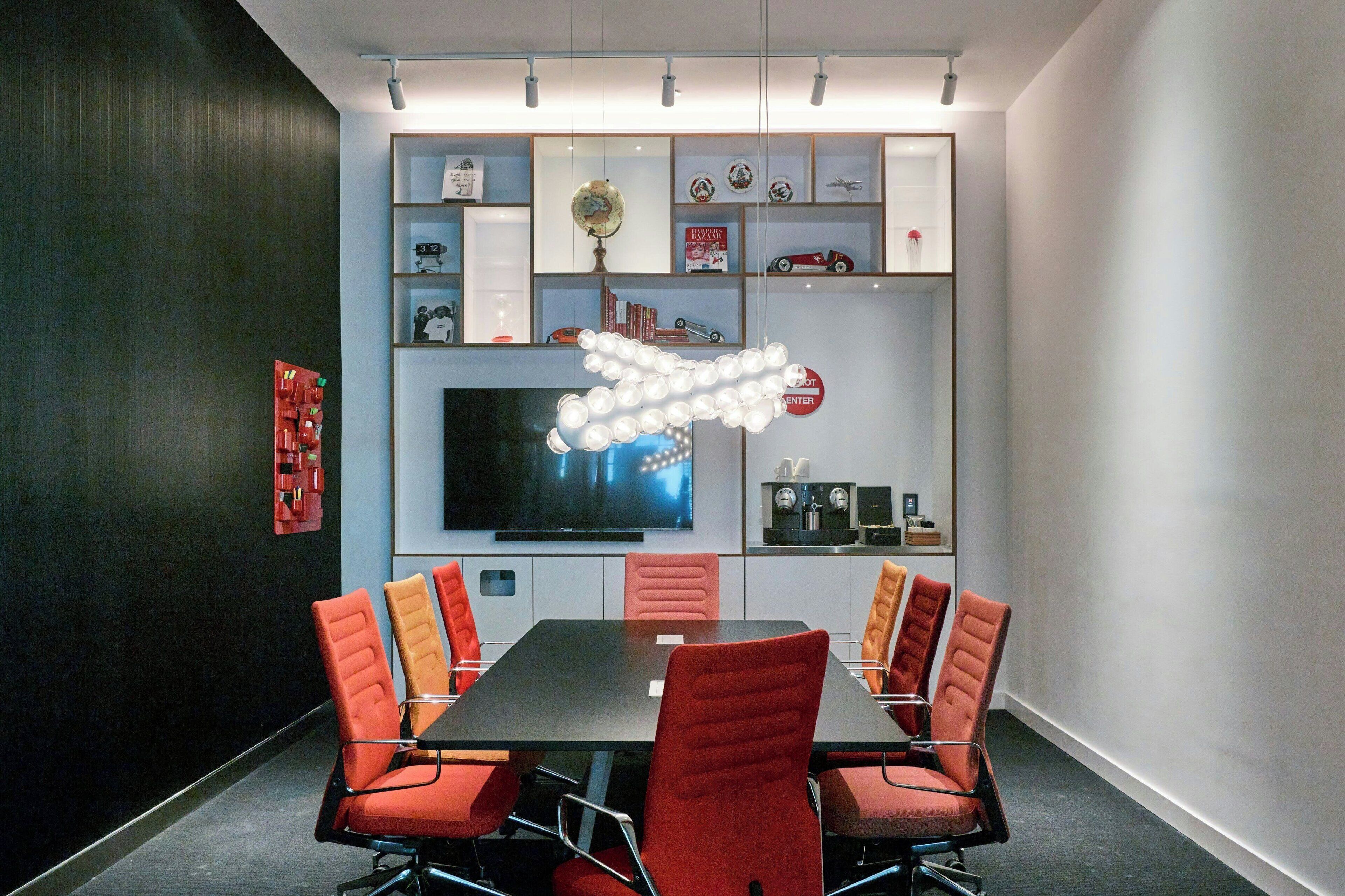 A conference room with a long table, red chairs, a flat-screen TV, and shelves with various items.