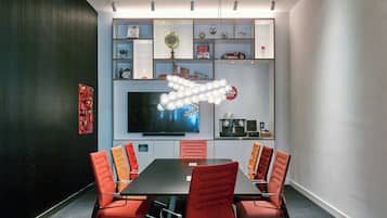 A conference room with a long table, red chairs, a flat-screen TV, and shelves with various items.