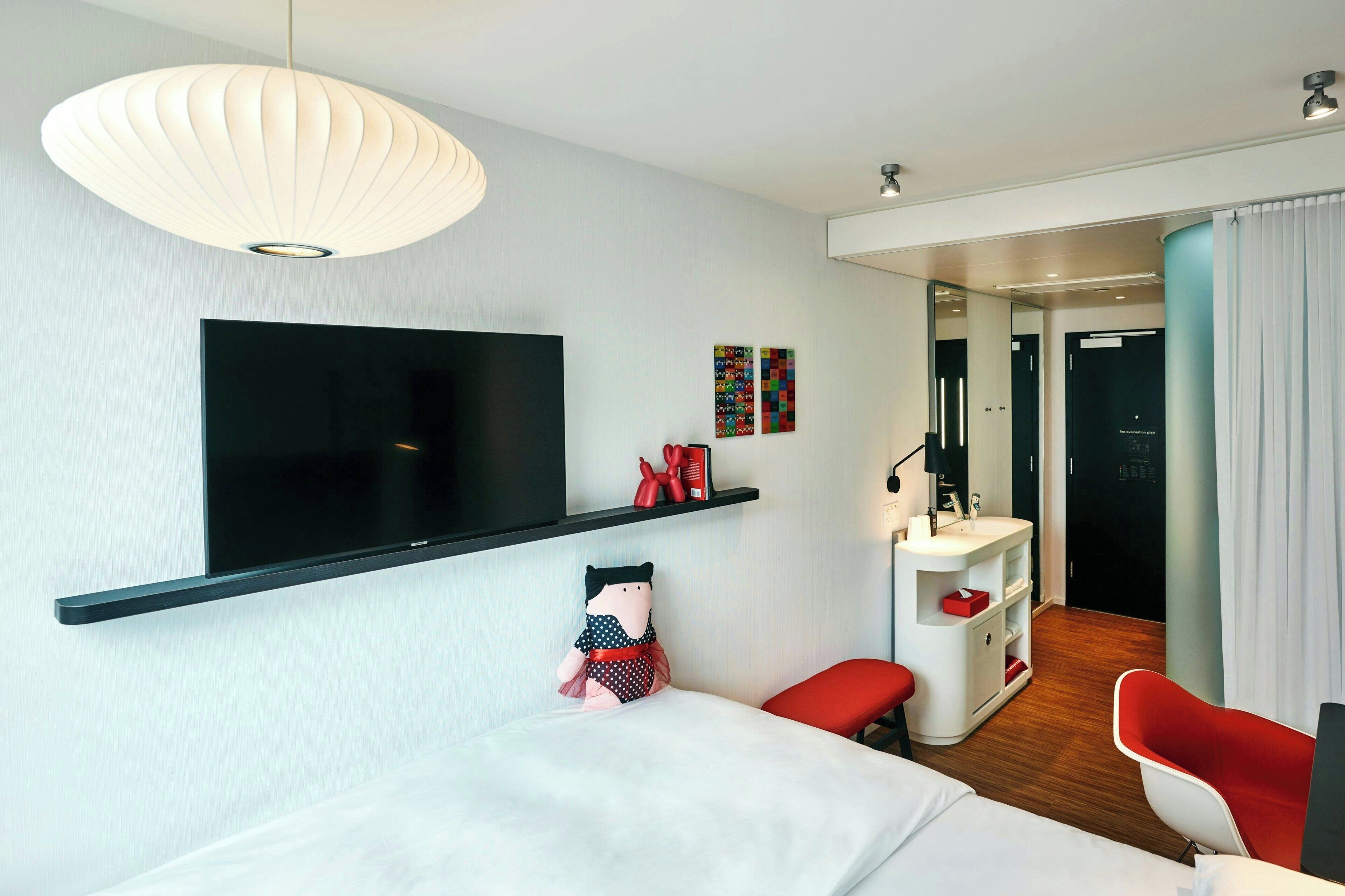 Et moderne hotelværelse med et fladskærms-tv, en seng, en rød lænestol, et hvidt skrivebord og et badeværelse, der kan ses gennem en åben dør.