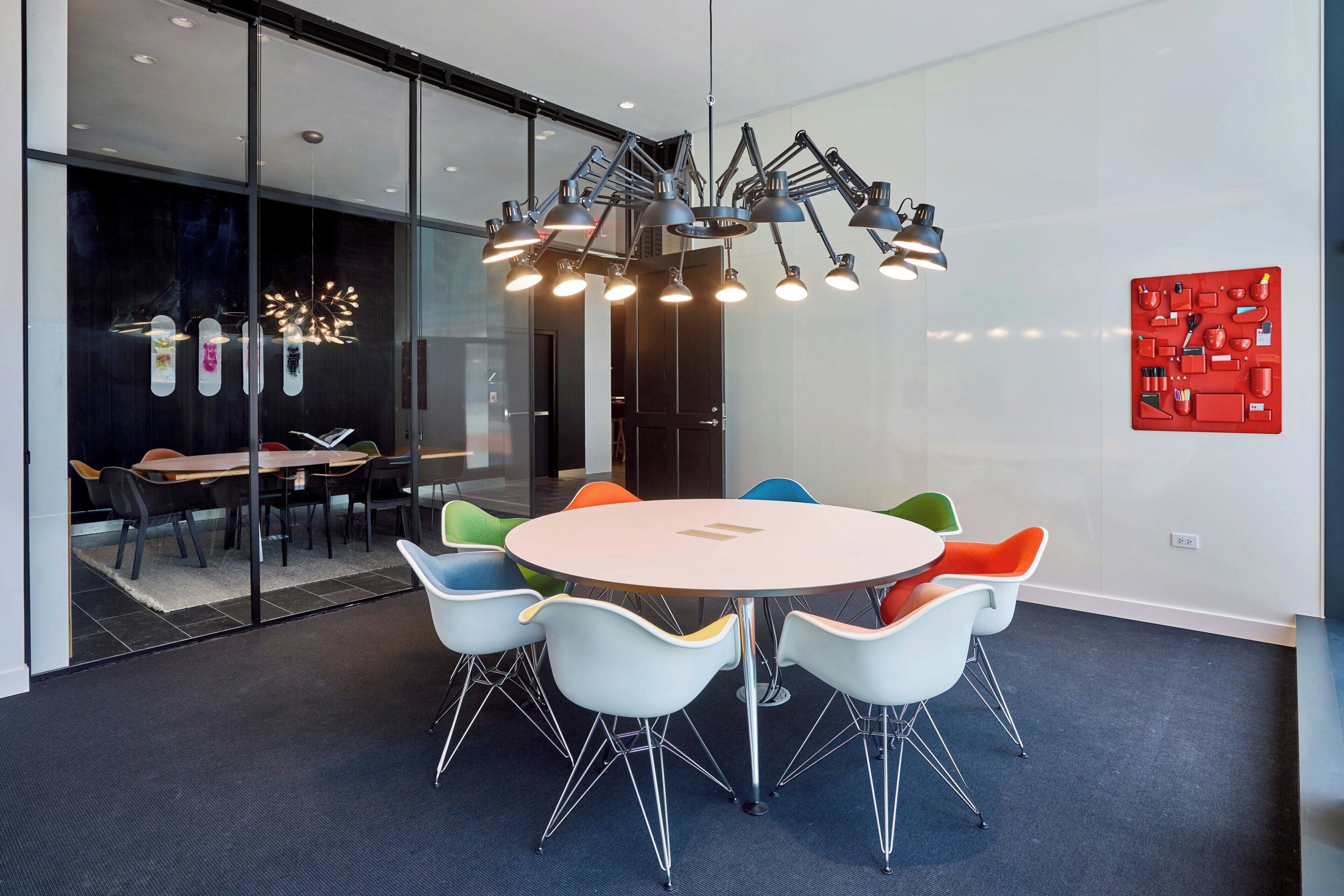 Een moderne vergaderruimte met een ronde tafel, kleurrijke stoelen en een bijzondere plafondlamp.